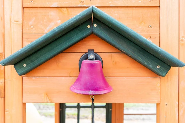 Pretend pink school bell on a wooden playground outdoors, with a string for making the ding dong noise, while kids participate in outdoor play.