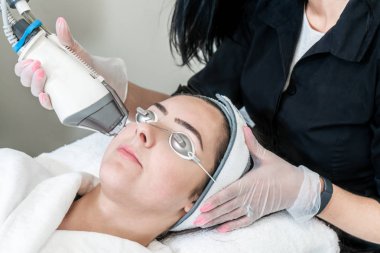 Tıbbi bir spa'da koruyucu gözlük takan bir kadın hastada akne izleri, kızarıklık ve kırışıklıkları gidermek için cilt gençleştirme yapan güzellik teknisyeni.