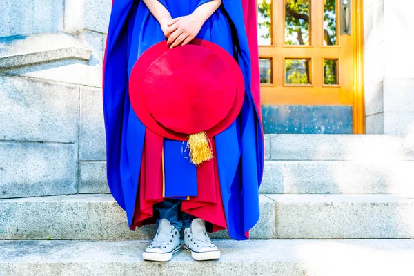 PhD (doctoral) graduate in regalia gown, holding Tudor bonnet cap, sitting on university steps, with sneaker canvas shoes showing. Red and blue grad gown, gold tassel showing.