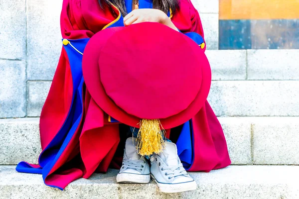 PhD (doctoraats) afgestudeerd in regalia Gown, holding Tudor Bonnet Cap, zittend op universitaire stappen, met sneaker canvas schoenen tonen. Rode en blauwe Grad Gown, gouden kwast tonen. — Stockfoto