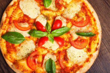 Üst görünüm, erimiş mozzarella peyniri, kırmızı kiraz domates ve taze yeşil fesleğen, mantar, kırmızı tatlı biber ve kiraz ile süslenmiş temiz bir kahverengi masada yaprakları ile Italyan pizza Margherita yakın