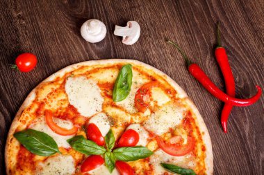 Üst görünüm, erimiş mozzarella peyniri, kırmızı kiraz domates ve taze yeşil fesleğen, mantar, kırmızı tatlı biber ve kiraz ile süslenmiş temiz bir kahverengi masada yaprakları ile Italyan pizza Margherita yakın