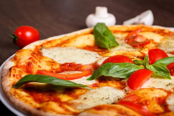 Primer plano margherita de pizza italiana con queso mozzarella derretido, tomates cherry rojos y hojas de albahaca verde fresca en una mesa marrón decorada con champiñones, pimiento dulce rojo y tomates cherry — Foto de Stock