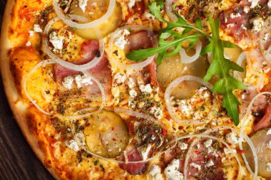 Yakın yukarı görünümü eritilmiş peynir ile İtalyan Pizza, mozzarella, jambon, turşu salatalık, soğan ve taze roka yaprakları kahverengi bir masada mantar, kırmızı tatlı biber ve kiraz domates ile süslenmiş