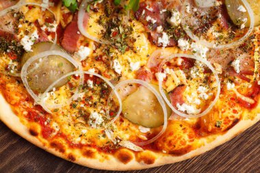 Yakın yukarı görünümü eritilmiş peynir ile İtalyan Pizza, mozzarella, jambon, turşu salatalık, soğan ve taze roka yaprakları kahverengi bir masada mantar, kırmızı tatlı biber ve kiraz domates ile süslenmiş