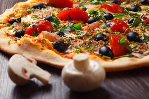 Pizza italiana de cerca con queso derretido, tomates rojos cherry, aceitunas negras, jamón y hojas verdes en una mesa marrón decorada con champiñones, chile rojo picante y tomates cherry — Foto de Stock