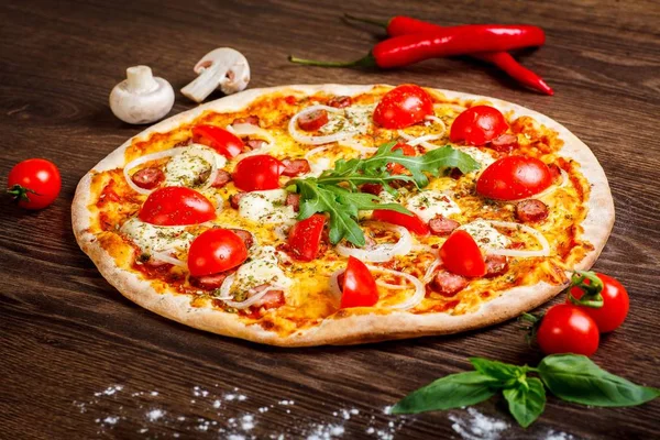 Pizza italiana con queso mozzarella derretido, tomates cherry rojos, salchichas y hojas de orégano verde fresco en una mesa marrón decorada con champiñones, pimiento dulce rojo y tomates cherry — Foto de Stock