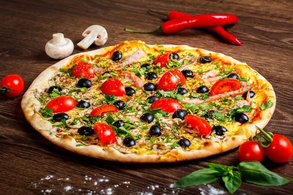 Pizza italiana con queso derretido, tomates rojos cherry, aceitunas negras, jamón y hojas verdes sobre una mesa marrón decorada con champiñones, pimiento rojo dulce y tomates cherry — Foto de Stock