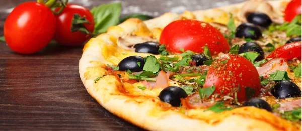 Närbild italiensk pizza med smält ost, röda körsbärstomater, svarta oliver, skinka och gröna blad på ett brunt bord dekorerad med svamp, Red Hot Chili Pepper och körsbärstomater — Stockfoto