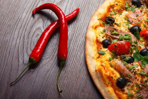 Pizza italiana de cerca con queso derretido, tomates rojos cherry, aceitunas negras, jamón y hojas verdes en una mesa marrón decorada con champiñones, chile rojo picante y tomates cherry — Foto de Stock