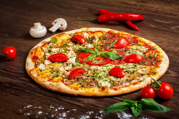 Close up Italian pizza with melted cheese, red cherry tomatoes, chicken, hen and fresh basil green leaves on a brown table decorated by mushrooms, red sweet pepper and cherry tomatoes — Stock Photo, Image