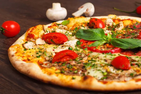 Närbild italiensk pizza med smält ost, röda körsbärstomater, kyckling, höna och färska basilika gröna blad på ett brunt bord dekorerad med svamp, röd paprika och körsbärstomater Stockbild