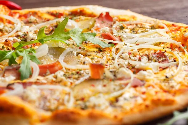 Närbild italiensk pizza med smält ost, mozzarella, skinka, inlagda gurkor, lök och färska ruccola blad på ett brunt bord dekorerad med svamp, röd paprika och körsbärstomater Stockfoto