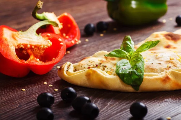 Närbild calzone italienska pizza med smält ost, tomater, färsk grön ört basilikablad på ett brunt bord dekorerad med svamp, svarta oliver, röd och grön peppar och rucola blad — Stockfoto