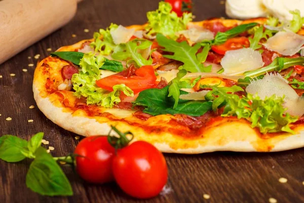 Close up pizza italiana con mozzarella, queso parmezano, tomates rojos y albahaca verde fresca, rúcula y hojas de col en una mesa marrón decorada con mozzarella, pimiento rojo, cereza y rodillo — Foto de Stock