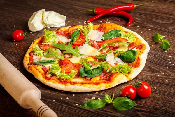 Close up pizza Italian with mozzarella, parmezan cheese, red tomatoes and fresh green basil, arugula and cabbage leaves on a brown table decorated by mozzarella, red pepper, cherry and rolling pin — Stock Photo, Image
