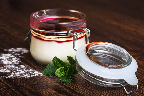 Delicious Italian dessert panna cotta with berry sauce in a glass jar on dark background decorated by sugar powder and fresh mint leaves — Stock Photo, Image