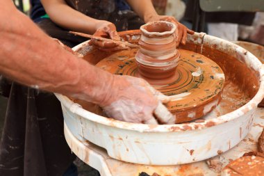 Çömlekçiliği kapatın. Yetişkin çömlekçi çamurlu elleri bir tekerlek üzerinde kil ile yardımcı olmak için çocuk elleri rehberlik. Çocuk basit bir kil pot veya plaka yapmaya çalışıyor