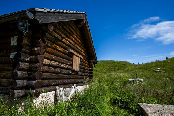 Wooden mountain cabin in Asiago surrounded by green meadow under clear blue sky. Concept of rural life, alpine tradition, eco tourism and rustic charm in the Italian Alps.