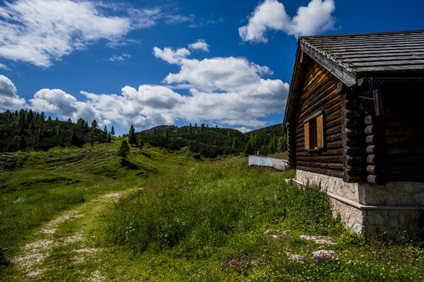 Wooden cabin in Asiago surrounded by alpine meadow and mountains under blue sky with clouds. Concept of rural life, eco tourism, mountain retreat and rustic charm in Italian Alps.