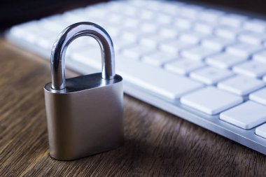 İle Metal asma kilit ve ahşap masa üstünde modern beyaz klavye yakın, hafif koyu tonu arka plan dim. Dijital veri koruma, şifreleme, internet erişimi, siber ağ, parola güvenlik kavramları.