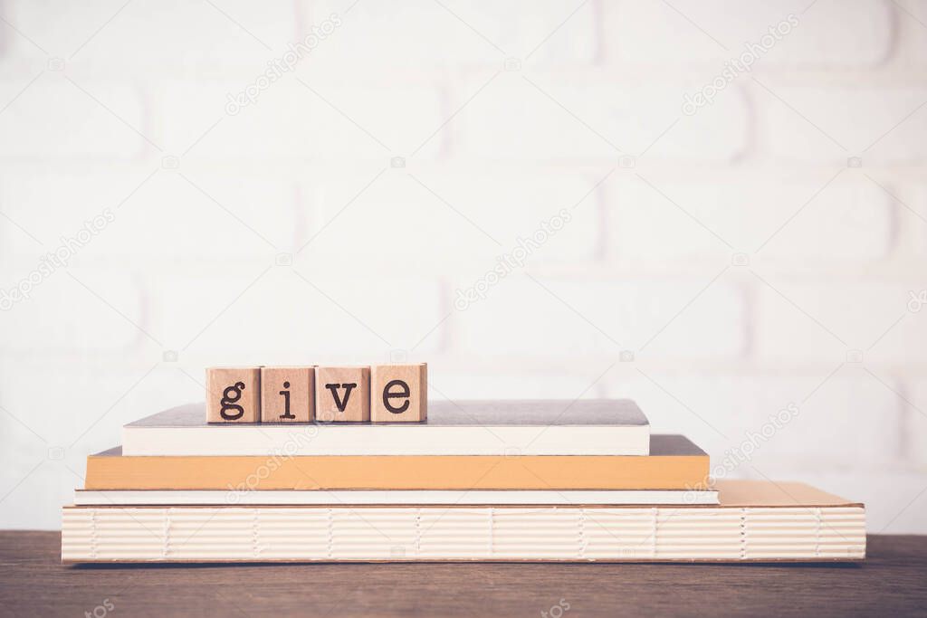 The word GIVE, letters on block cubes on top of books and wooden table with white bricks background, blank copy space, vintage minimal style. Concepts of share, charity, donation and love.