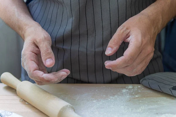 The hands of the chef knead the dough on a wooden table near the milling molds for ravioli rolling pin on a gray background. The cooking process
