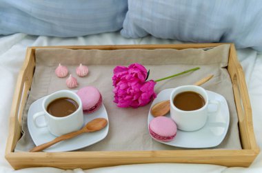 İki kişilik yatakta kahvaltı. Kahve, makaron ve Bizet ile ahşap tepsi. Dekorasyon pembe şakayık. Pencereden güzel doğal ışık.