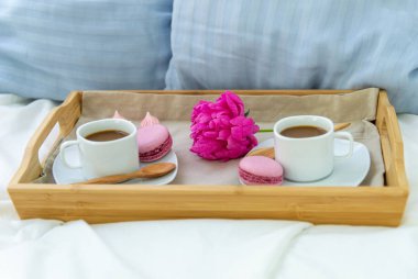 İki kişilik yatakta kahvaltı. Kahve, makaron ve Bizet ile ahşap tepsi. Dekorasyon pembe şakayık. Pencereden güzel doğal ışık.