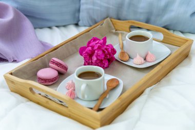 İki kişilik yatakta kahvaltı. Kahve, makaron ve Bizet ile ahşap tepsi. Dekorasyon pembe şakayık. Pencereden güzel doğal ışık.