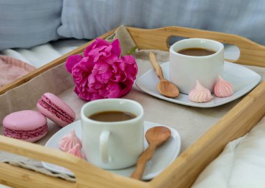 İki kişilik yatakta kahvaltı. Kahve, makaron ve Bizet ile ahşap tepsi. Dekorasyon pembe şakayık. Pencereden güzel doğal ışık.