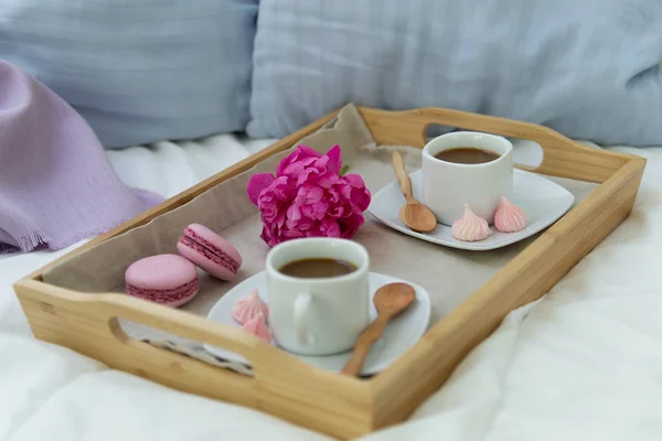 Breakfast in bed for two. Wooden tray with coffee, macaroons and Bizet. Decoration pink peony. Beautiful natural light from the window.