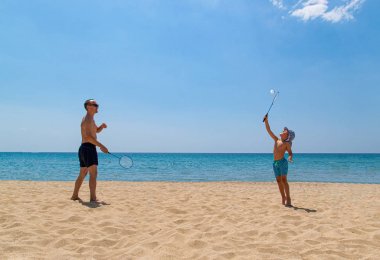 Baba ve oğul sahilde badminton raket ve shuttlecock ile oynamak. Aile tatili konsepti. Sahilde parlak güneşli bir gün.