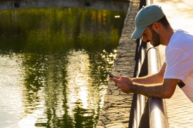 Genç adam, dirsekleri göldeki korkuluklara bağlı bir şekilde cep telefonuna bakıyor.