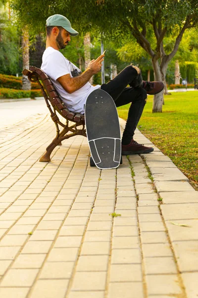 Młody człowiek siedzi na ławce patrząc na telefon komórkowy z łyżwy pionowe linie bruk — Zdjęcie stockowe