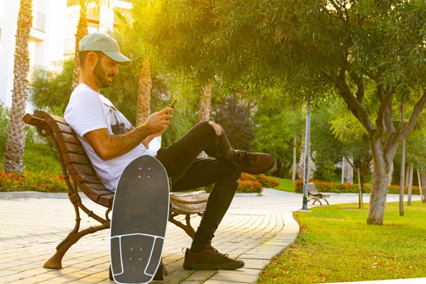 Młody człowiek siedzi na ławce patrząc na telefon komórkowy z łyżwy ze światłem zachodzącego słońca — Zdjęcie stockowe