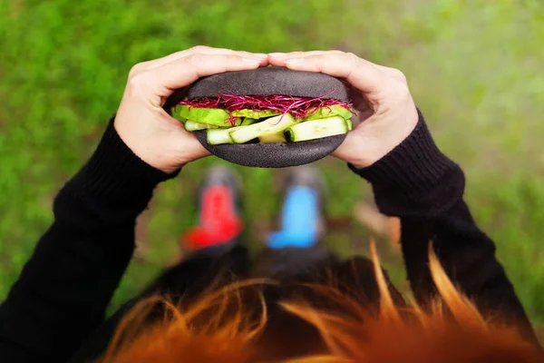 Beautiful woman eating healthy black bun fresh vegetable burger outdoors in nature.