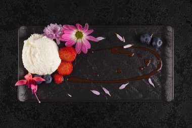 Lezzetli vanilyalı dondurma yenilebilir çiçekler ve koyu arka plan üzerinde siyah plaka üzerinde taze meyve ile devam eder. Düz yatıyordu.