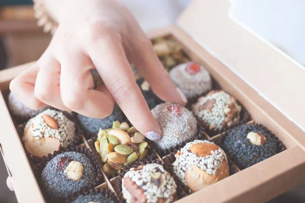 The box with healthy organic sweets with dried fruits, nuts, banana, dates and honey - vegan raw snack. Female hand takes a vegan candy.