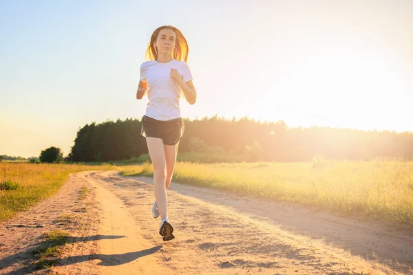 Young fitness girl in a white t-shirt running on the dirt road across the field in the sunrise.