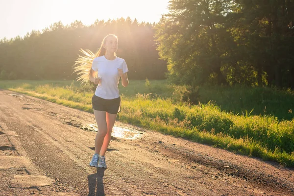 Off-road running. Young fitness girl in a white t-shirt running on the dirt road across the field in the sunrise.