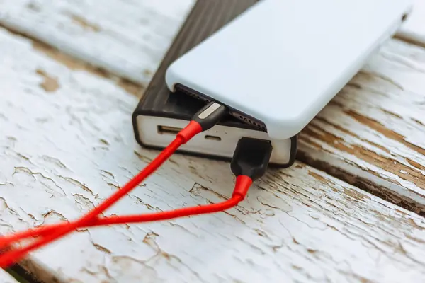 stock image Phone charging with energy bank and a red cable on an old, white, wooden table.