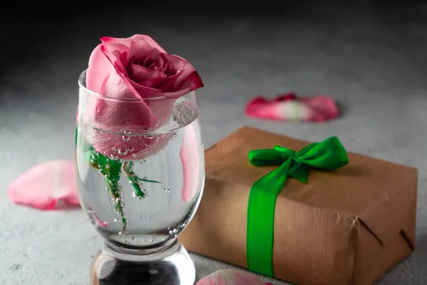 Rosa rosa en un vaso de agua, pétalos de rosa y una caja con un regalo en la mesa — Foto de Stock