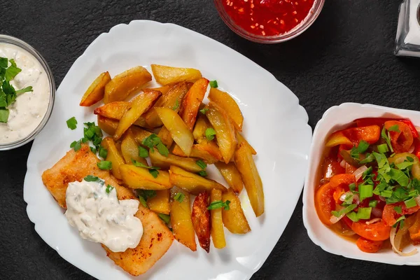 Pescado frito y patatas fritas en un plato blanco en negro la mesa de la cocina con salsa de tomate, salsa tártara y ensalada de verduras en vinagre - foto, imagen — Foto de Stock