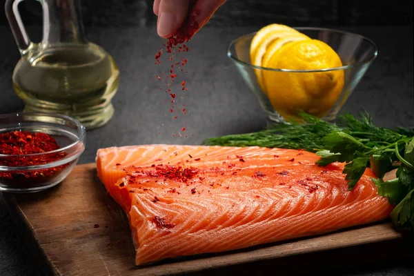 Female hand sprinkles a piece of salmon fillet lying on a wooden cutting board with spices - photo, image — Stock Photo, Image