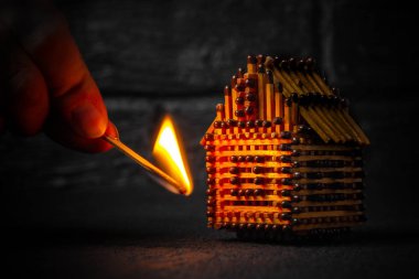 hand with a burning match sets fire to the house model of matches, risk, property Insurance protection or ignition of combustible materials concept clipart
