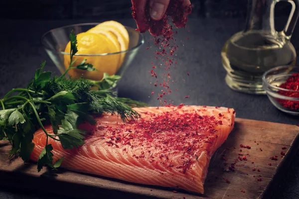 Female hand sprinkles a piece of salmon fillet lying on a wooden cutting board with spices - photo, image — Stock Photo, Image