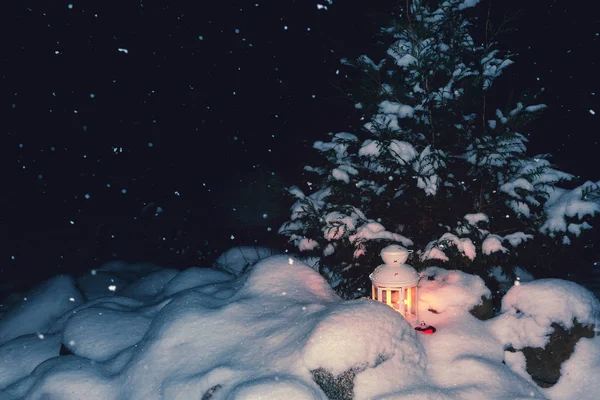 Lykta med ett brinnande ljus under en snötäckt julgran på gården i huset i snödrivorna — Stockfoto