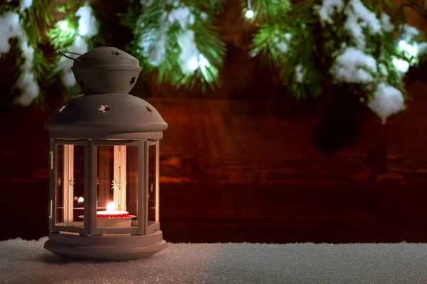 Lanterna con una candela accesa sulla neve sullo sfondo di una vecchia parete di legno decorata con rami di albero di Natale e luci di Natale. Copia spazio per testo — Foto Stock