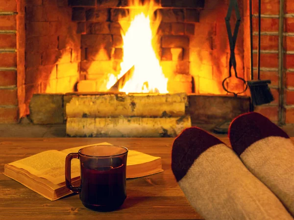 Tee, Buch, Frauenfüße in warmen Socken auf einem Holztisch vor einem brennenden Kamin — Stockfoto